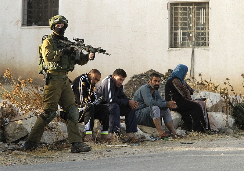 An Israeli soldier keeps watch as Palestinians sit nearby after the army entered the village of Yatta in the occupied West Bank on June 9,2016 (AFP)